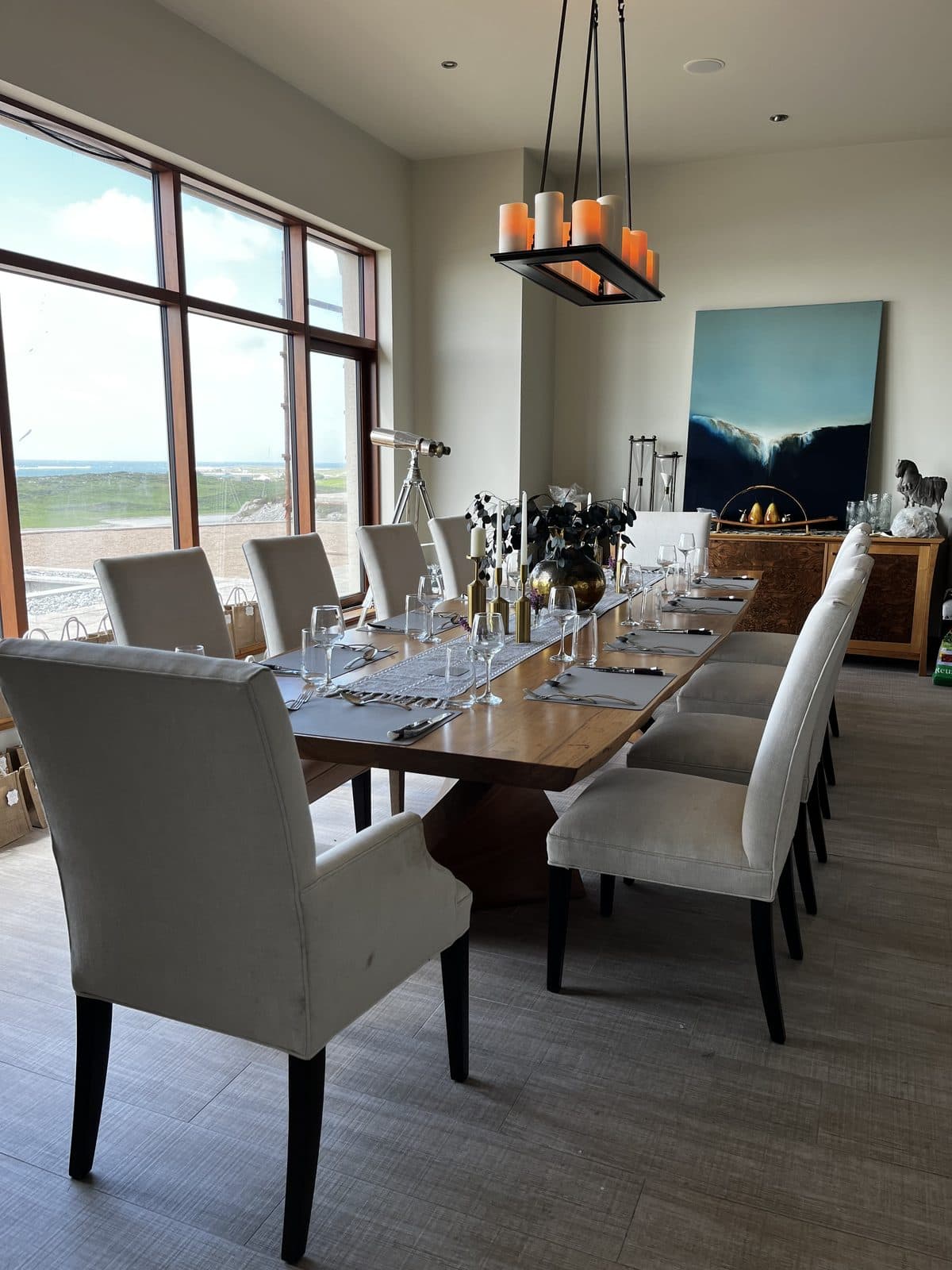 A long wooden dining table dressed for service in a country-house dining room — linen runner, glassware, candle centrepiece, brass and silver bottles, linen-upholstered chairs along both sides, an oversized window on the left looking out over the Atlantic coastline, and a modern chandelier of orange candles overhead.