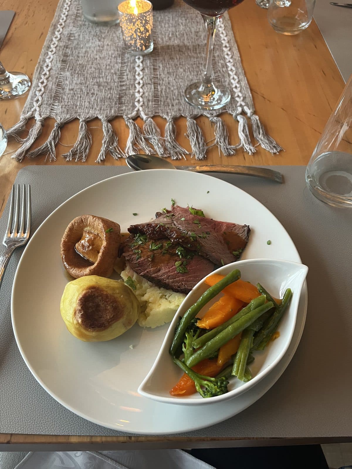 An overhead shot of a plated dinner — sliced roast beef with fresh herbs, mashed potato, a Yorkshire pudding, and mixed vegetables in a curved white serving dish, set on a wooden table with a fringed linen runner, wine glass, and tealight.