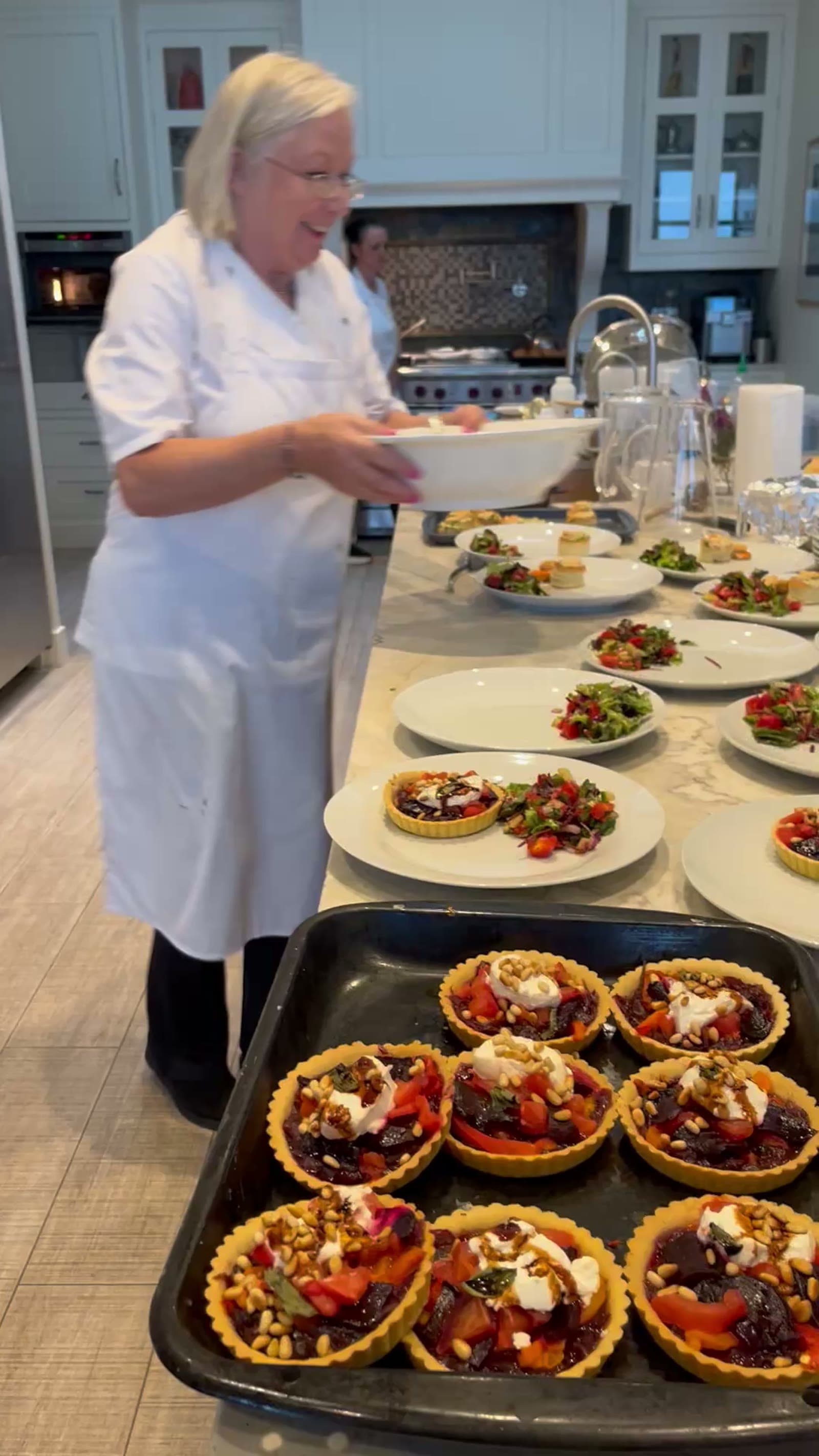 Dóirín in chef whites mid-plating in the venue kitchen, smiling, holding a white serving bowl, with a tray of beetroot-and-goats-cheese tarts in the foreground and plates of green salad receding down the marble counter behind her.