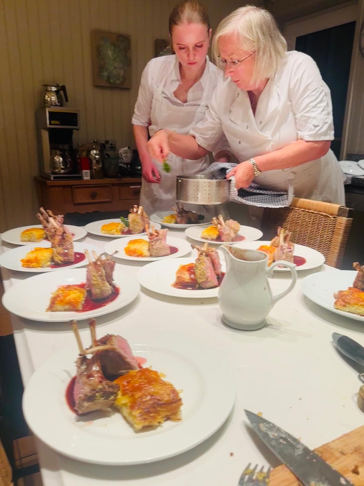 Two chefs in whites plating a row of rack-of-lamb plates finished with red-wine jus at a kitchen table.