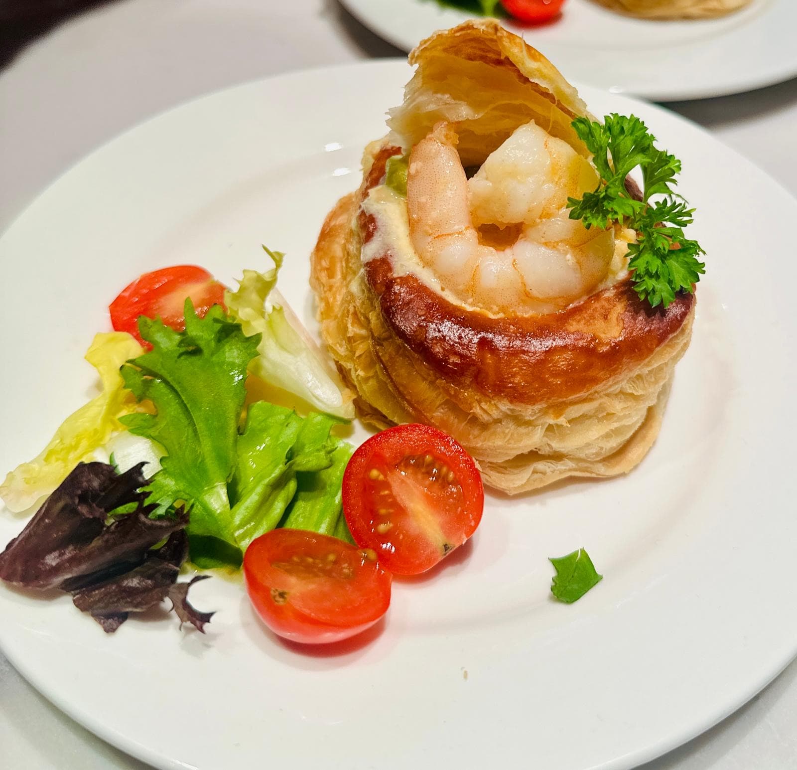 A single prawn vol-au-vent starter — golden puff pastry filled with prawns in a creamy sauce, topped with parsley, served with a small dressed salad and cherry tomatoes on a white plate.